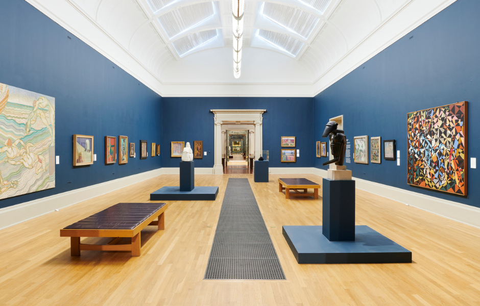 Tate Britain unveiled a complete rehang of its free collection displays in May 2023. Image of a long museum gallery with dark blue walls, a white domed ceiling, and bright modern paintings on either side.