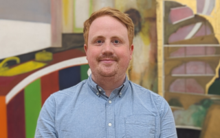 Jacob Weber, Collection Systems Manager, at Tate stands in front of an abstract painting. Jacob has short ginger hair, light facial hair, and blue eyes. He is smiling slightly and wearing a light blue button-up shirt.