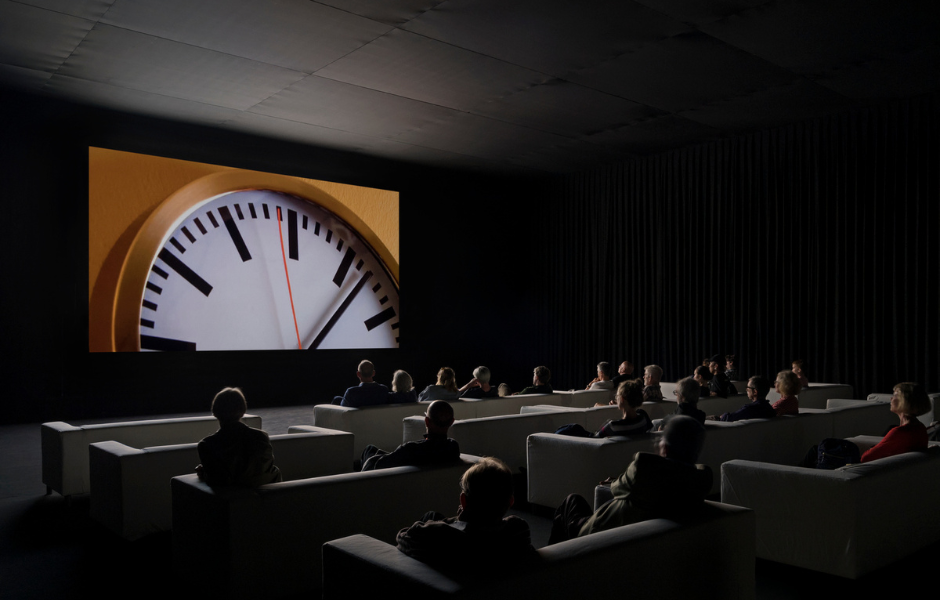 Photo of visitors at Tate Modern observing Christian Marclay's The Clock, 2010. A 24-hour video projection made from fragments of cinema footage where time is featured.